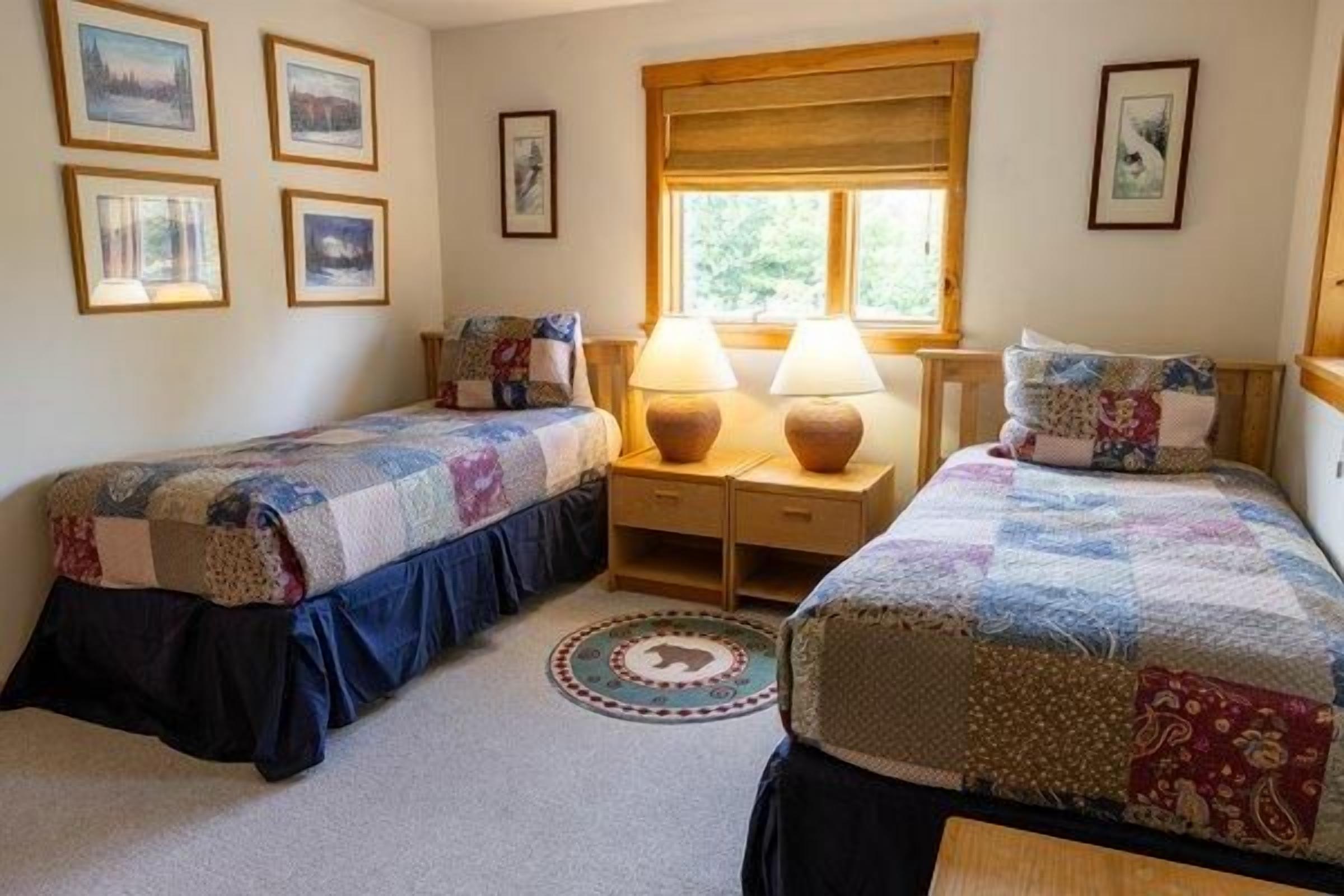A bedroom with two single beds, a window with blinds, a desk with lamps, and a rug with a circular design.