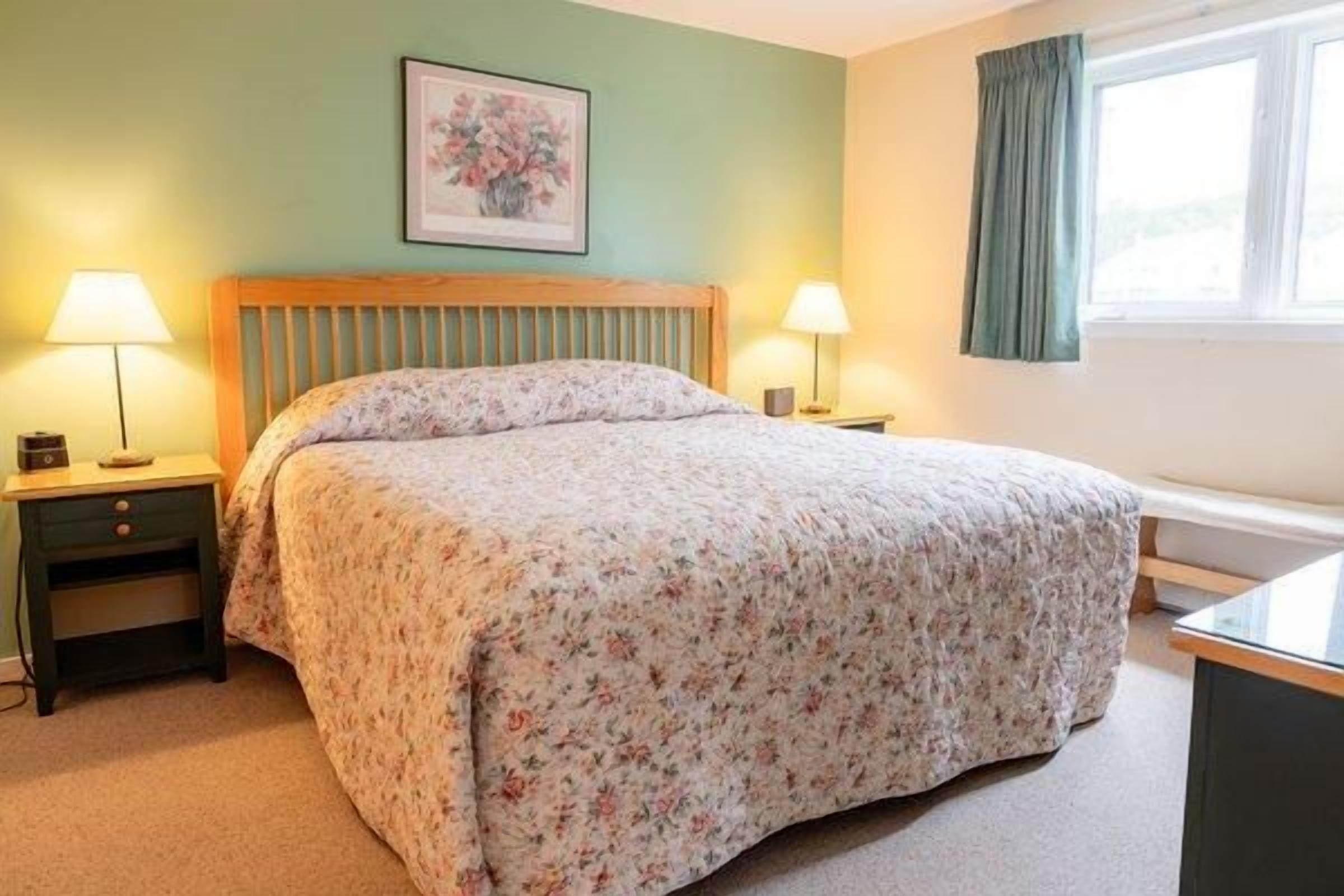 Un dormitorio con una cama, dos mesitas de noche con lámparas, una ventana con cortinas y un cuadro en la pared.
