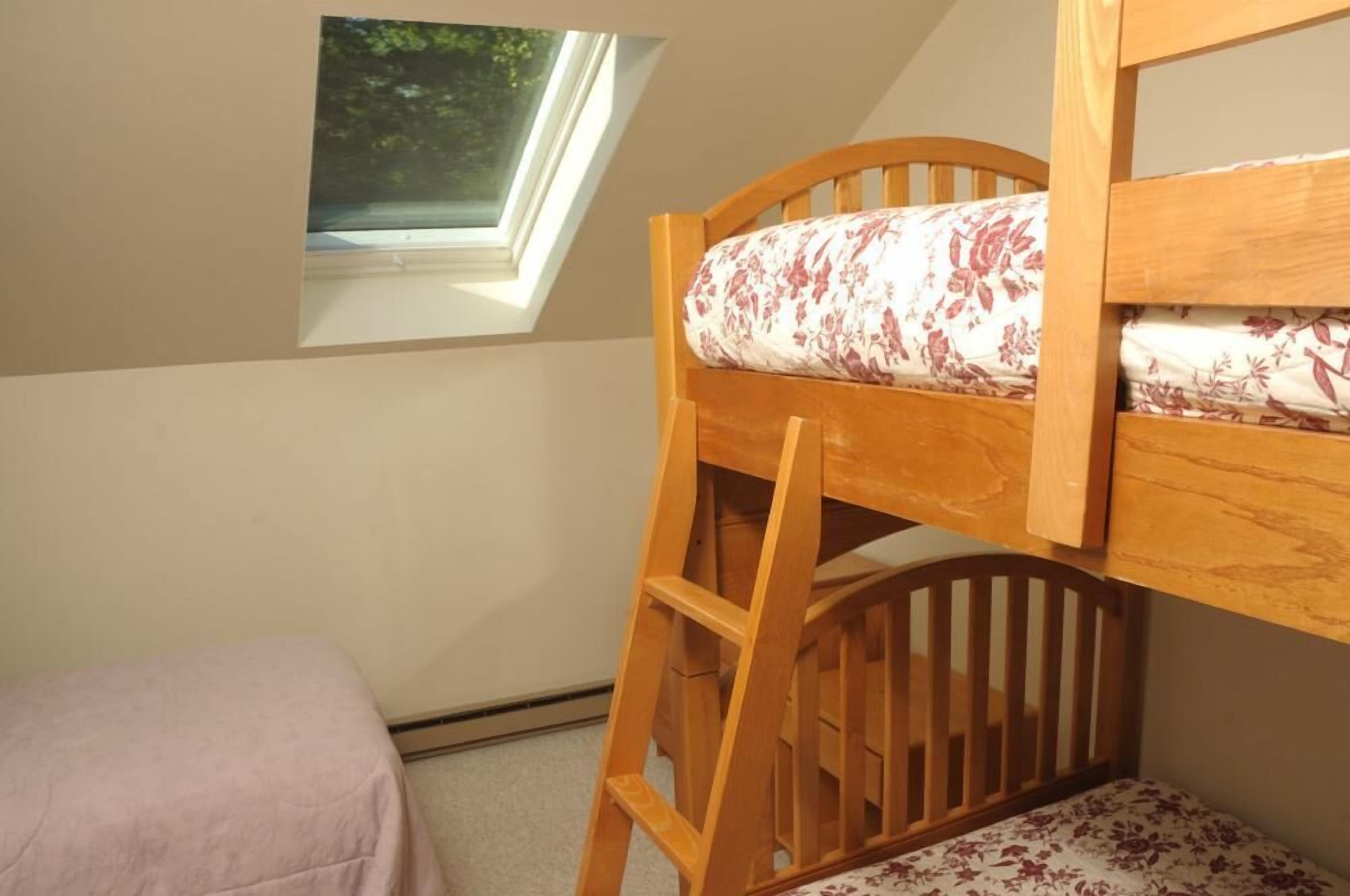 Una cama litera con escalera de madera, una ventana con vistas a la vegetación y una mesita de noche con un cojín estampado con motivos florales.