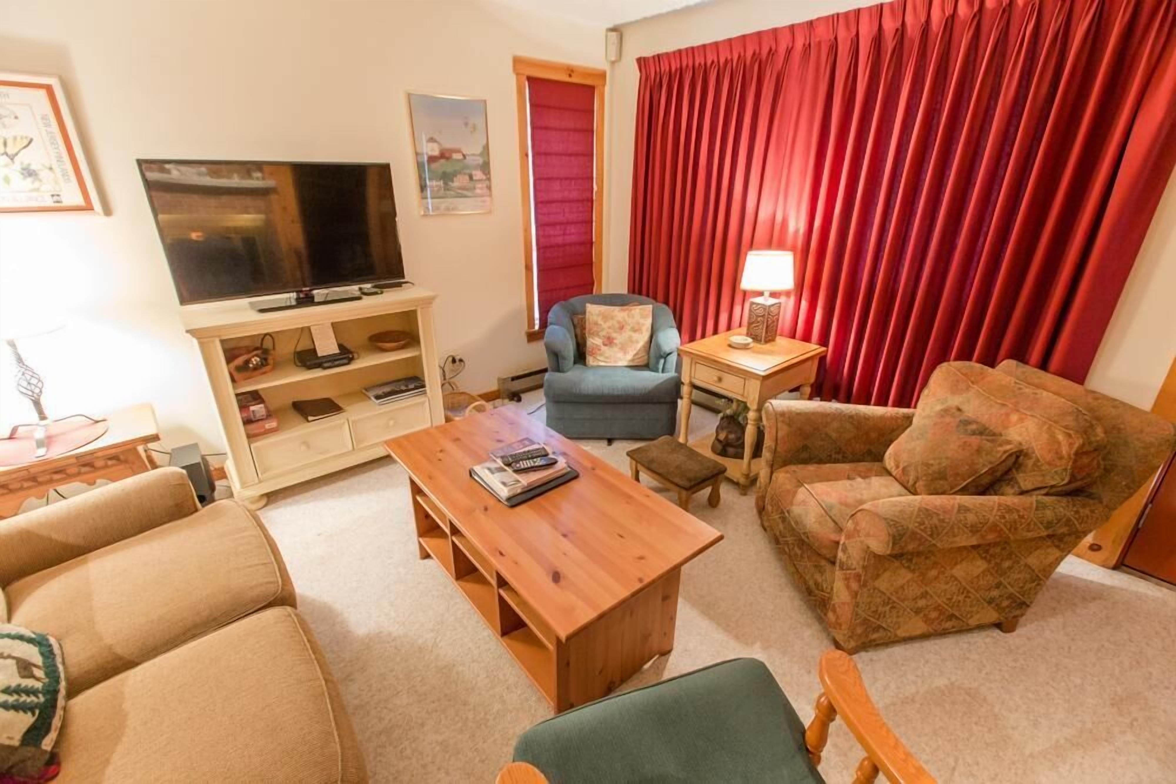 Una sala de estar con televisor, sofá, sillón, mesa de centro y cortina roja.
