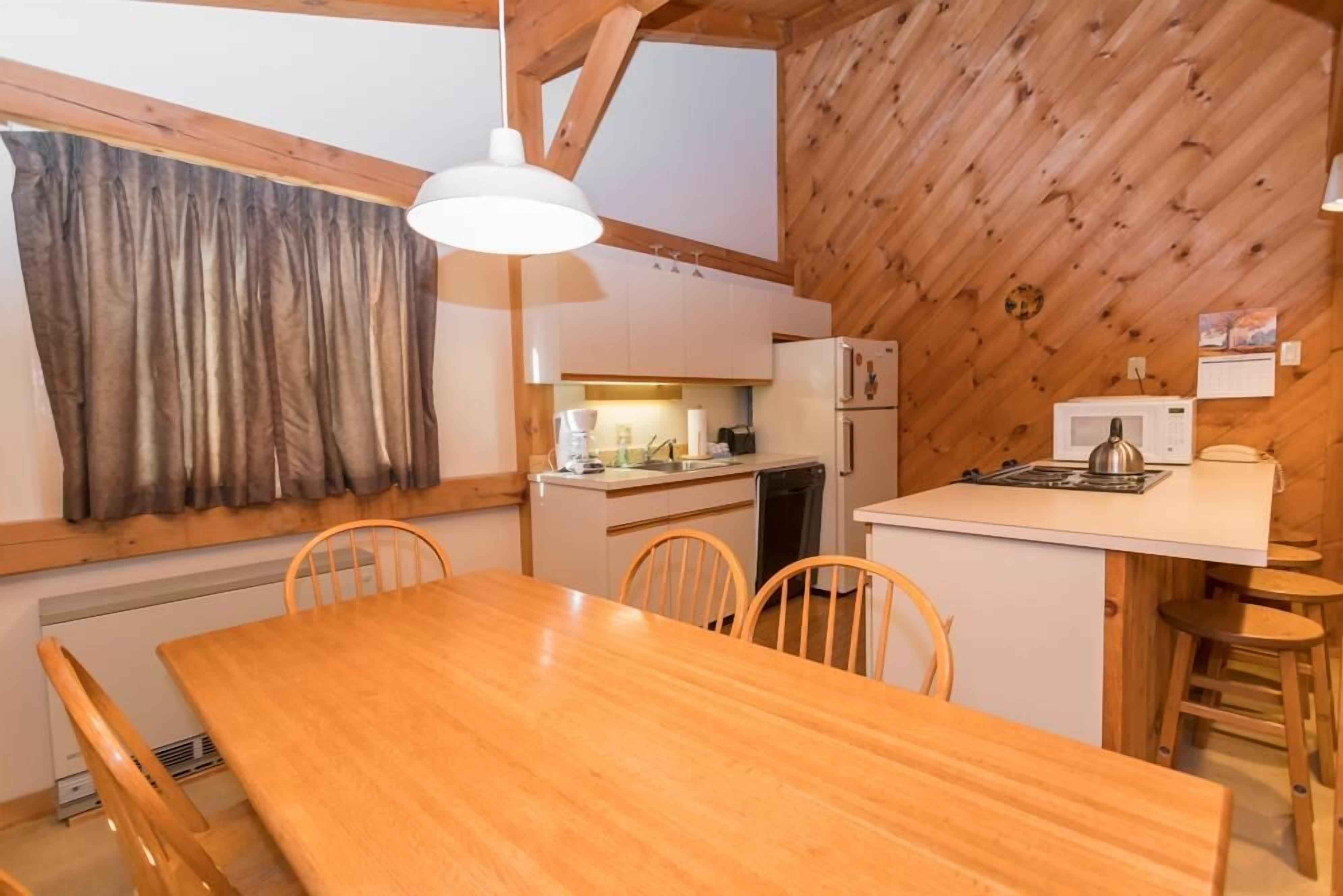 A wooden interior with a dining table, chairs, a kitchen area, and a window with curtains.
