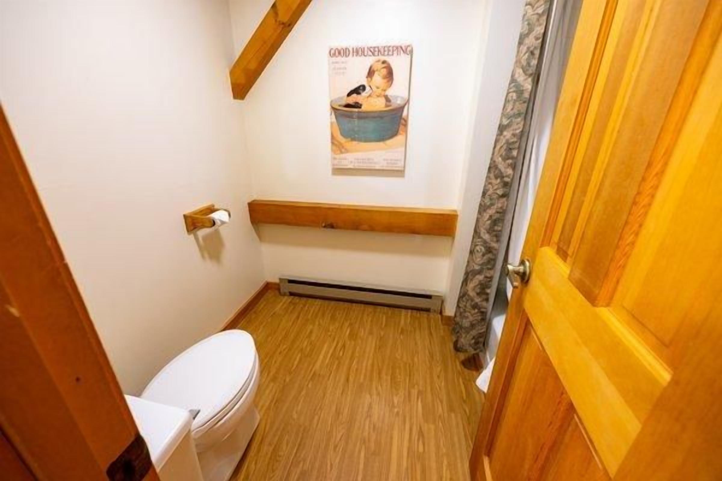 A small bathroom with a wooden floor, a white toilet, a wooden bench, and a poster on the wall.