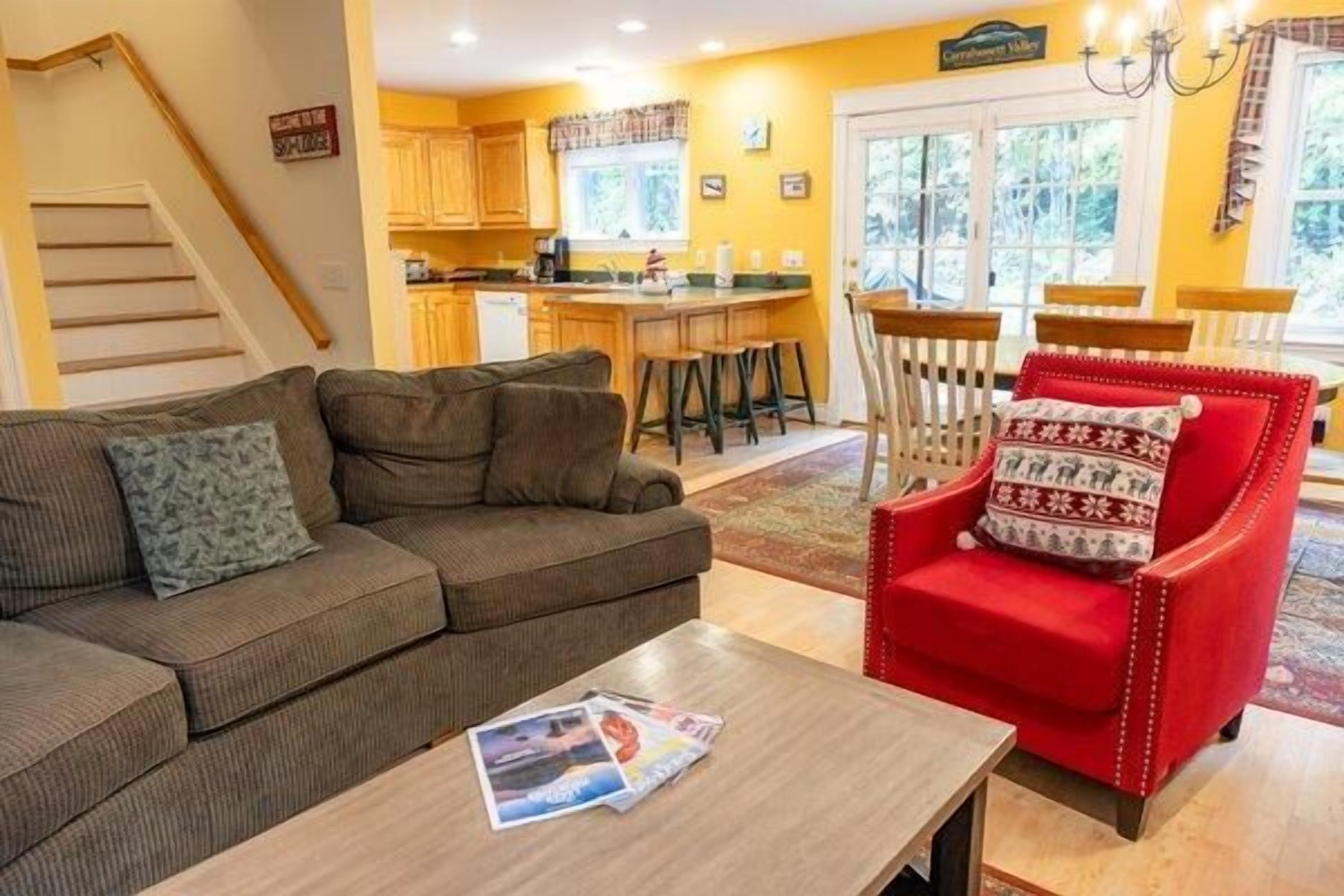 A living room with a sofa, a red armchair, and a coffee table. There is a staircase to the left and a kitchen in the background.