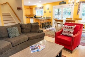 Un salon avec un canapé, un fauteuil rouge et une table basse. Un escalier se trouve à gauche, et une cuisine est visible au fond.