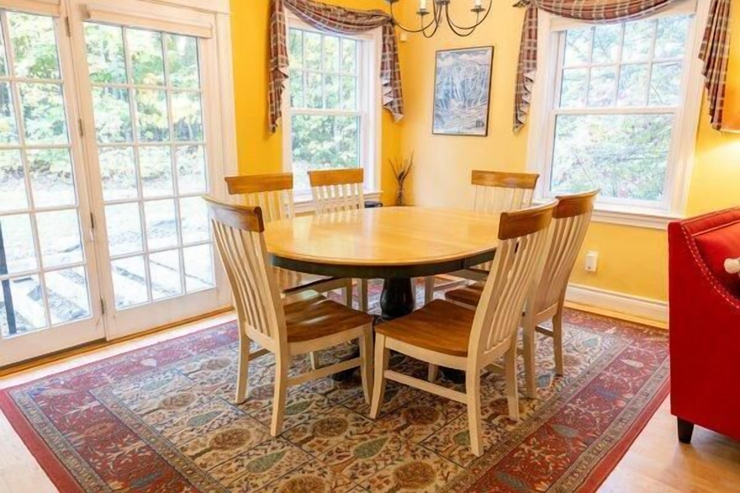 A dining area with a round table and six chairs, large windows with curtains, and a red chair in the corner.