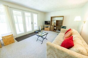 Ein Wohnzimmer mit einer Couch, einem Fernseher, einem Couchtisch und einer Glasschiebetür, die zu einem Außenbereich führt.