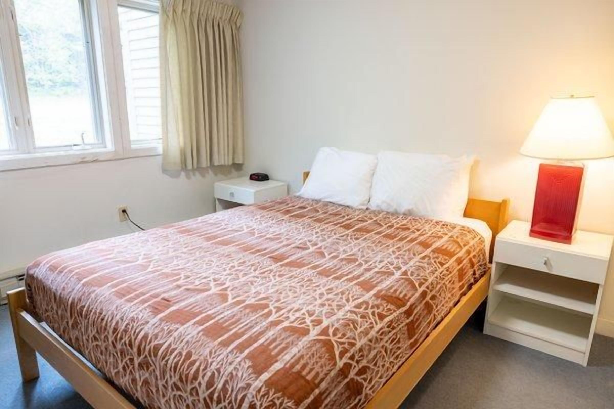 A neatly made bed with a patterned bedspread, a bedside table with a lamp, and a window with curtains.