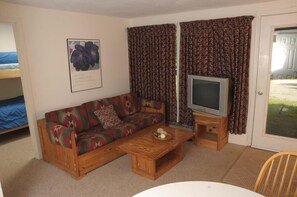 Ein möblierter Raum mit einer Couch, einem Fernseher, einem kleinen Tisch und einem Fenster mit Vorhängen.