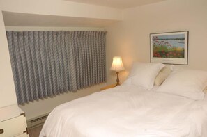 Ein Hotelzimmer mit Bett, Lampe, einem fenster mit Vorhängen und einem gerahmten Bild an der Wand.