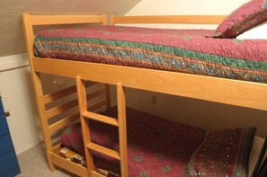 Ein Stockbett mit Holzrahmen, einer Matratze in Rot und Grün gemustert und einem Kopfkissen.