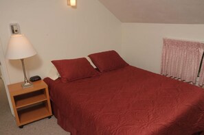A single bed with a red bedspread and two pillows, a nightstand with a lamp, and a radiator with pink curtains. - Sugarloaf Mountain Kibby Mountain Road C2315 (Carrabassett Vly)