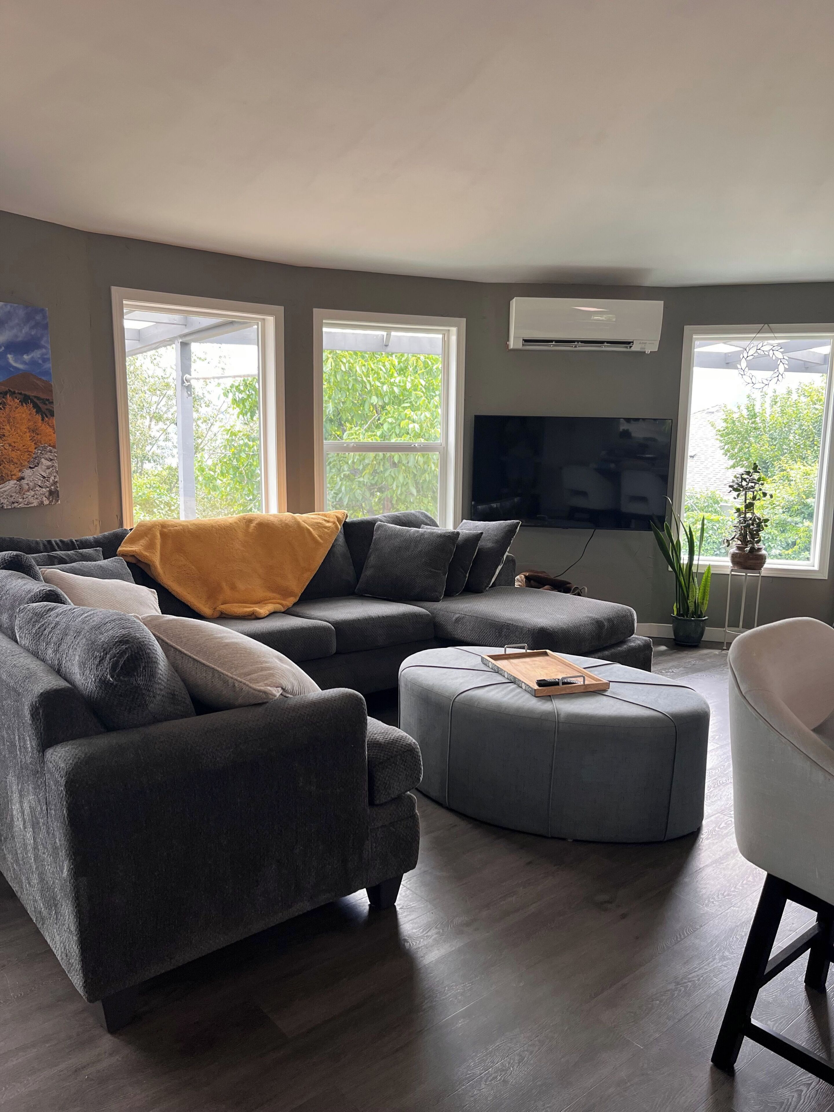 A modern living room with a sectional sofa, ottoman, and a white armchair. There are large windows, a flat-screen TV, and a potted plant.