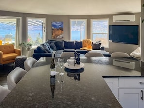 A modern living room with a large L-shaped sofa, a dining table with a bottle and glasses, and a TV mounted on the wall.
