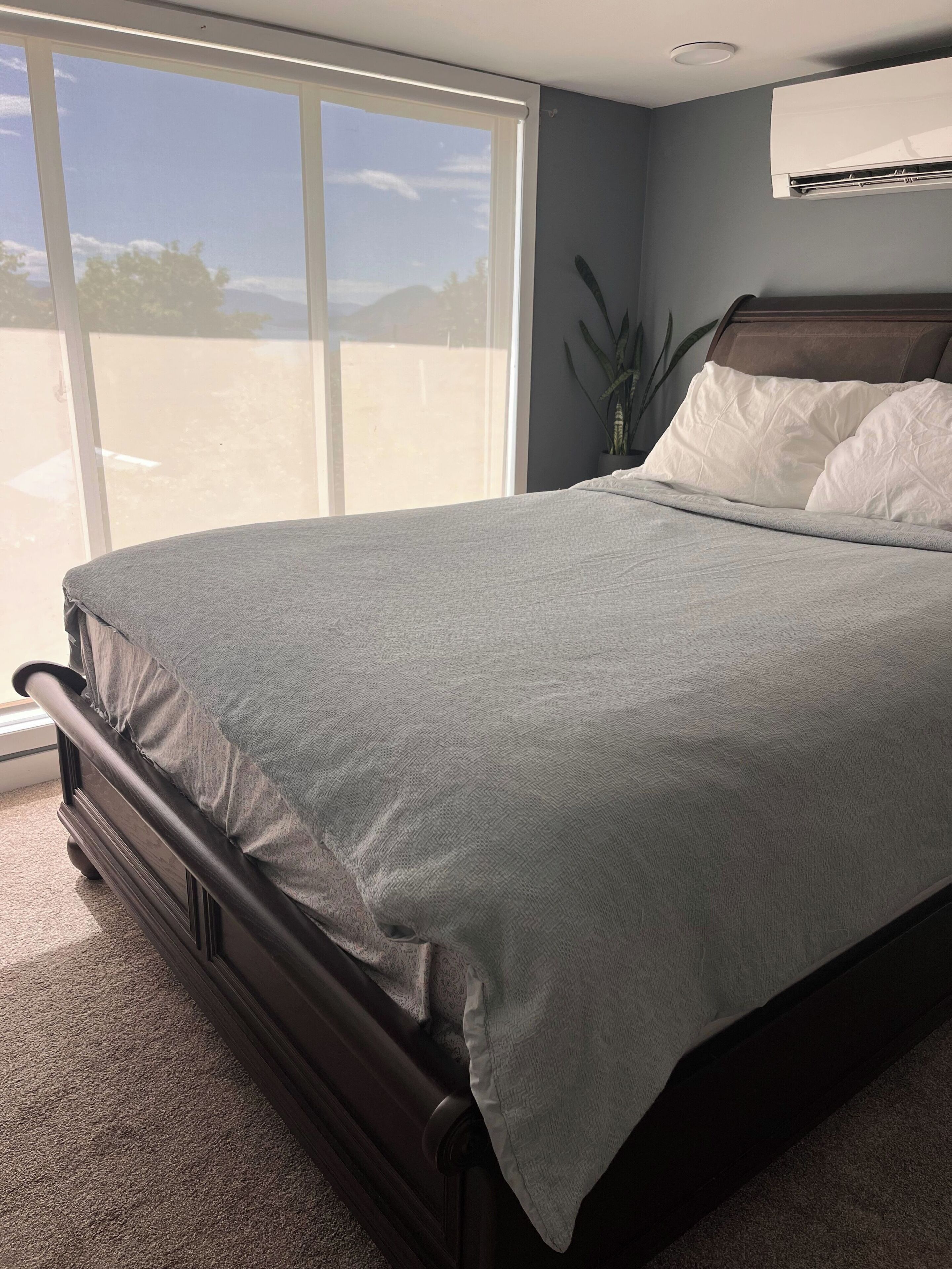 A bedroom with a large window, a bed with a grey bedspread, a wooden headboard, and a potted plant.