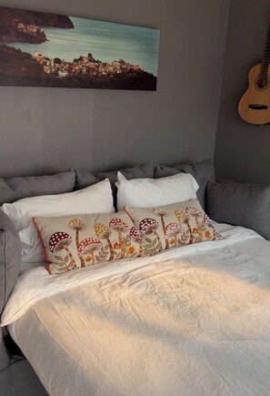 A bedroom with a bed, pillows, a guitar, and a painting of a coastal town.