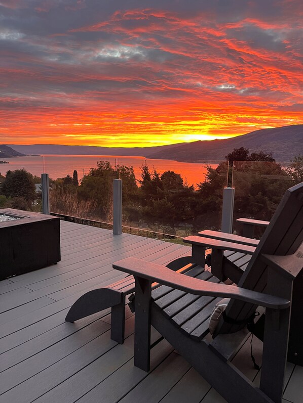 A deck with chairs and a fire pit overlooking a lake at sunset.