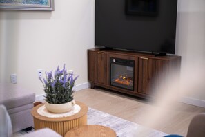 A modern living room with a flat-screen TV mounted on the wall, a wood-burning stove, a grey sofa, and a small round wooden table with a plant. - Luxurious 2BR Suite Near Langley Events Centre | King Bed | Fully Equipped (Langley Township)