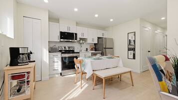 A modern kitchen with white cabinets, a microwave, stove, and refrigerator. There is a dining table with chairs and a bench.