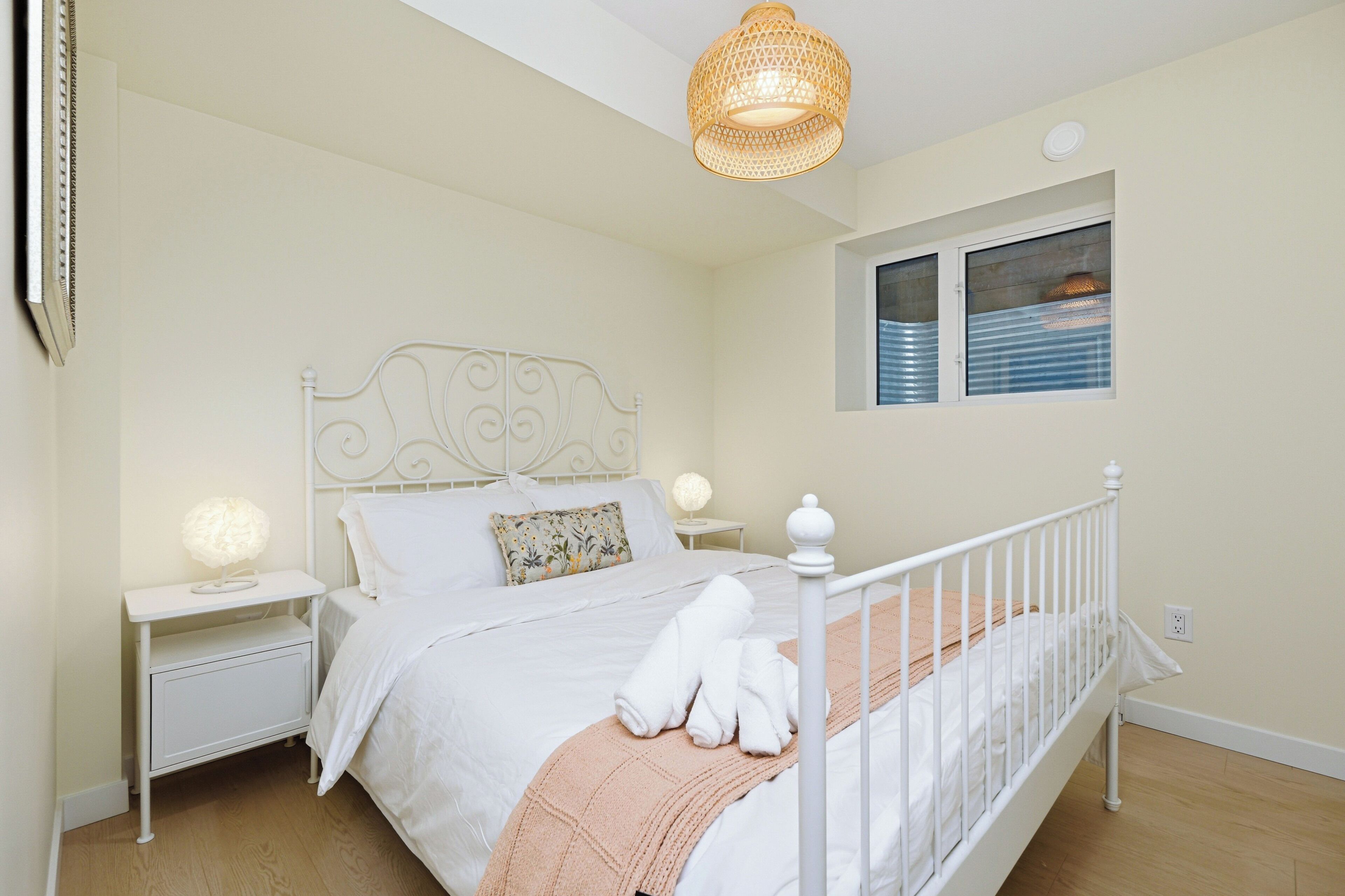 A bedroom with a white metal bed frame, a bedside table with a lamp, a window with blinds, and a hanging light fixture.