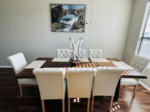 A dining room with a wooden table, white cushioned chairs, and a framed picture on the wall.