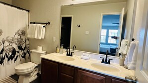 A bathroom with a double sink vanity, a toilet, a shower curtain with a floral design, and a large mirror with lights above it. - Spacious Luxe Retreat for family and groups Austin (Round Rock)