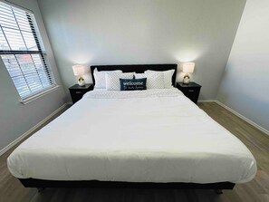 A neatly made bed with a 'welcome to our home' pillow, two bedside lamps, and a window with blinds.