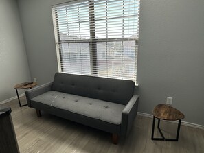 A room with a grey sofa, a small round table, and a window with blinds.