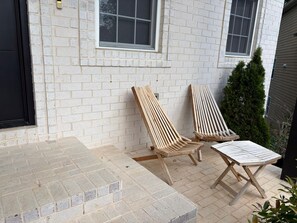 A patio with two wooden lounge chairs and a table, a white brick wall, and a window.