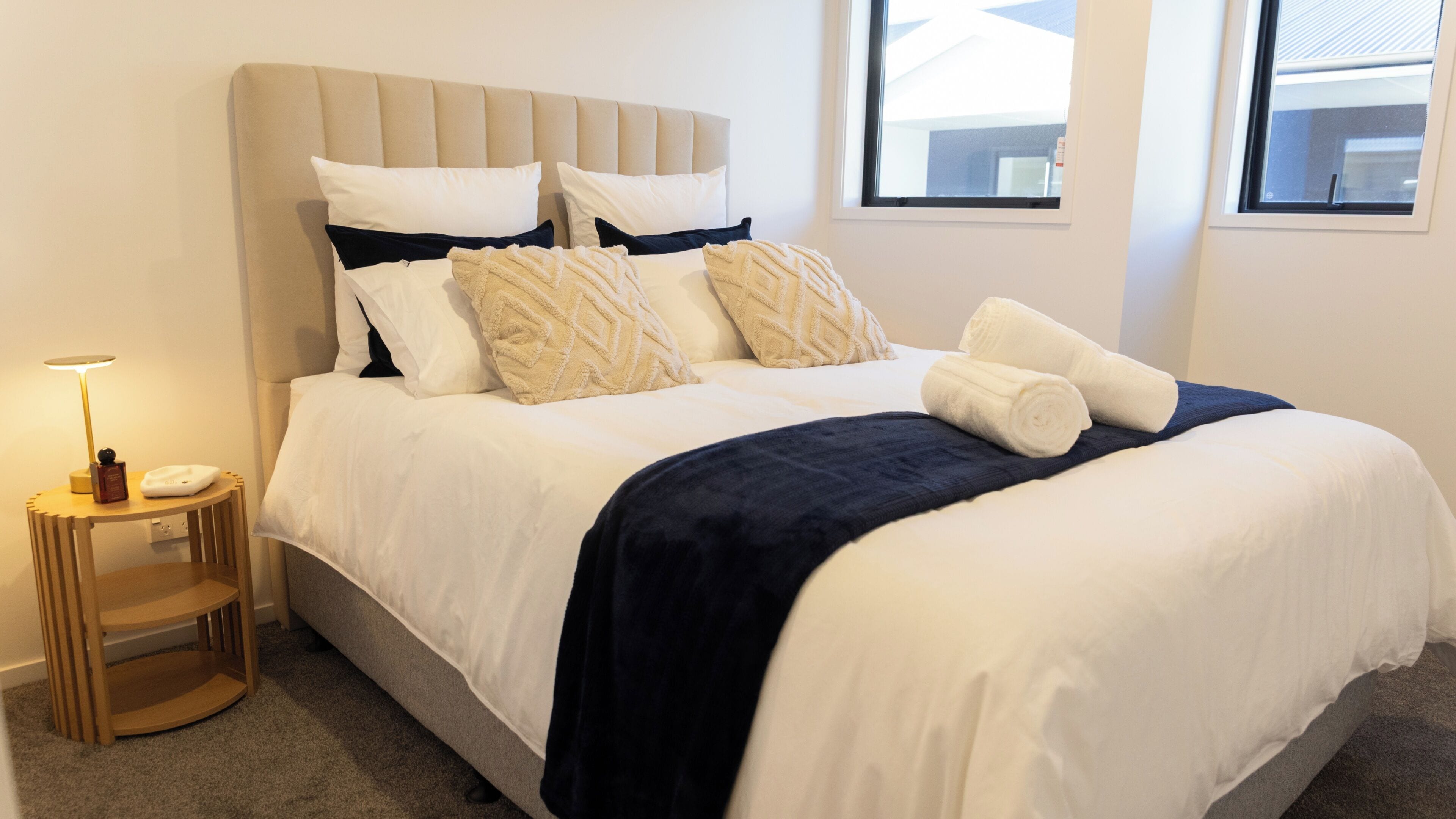 A neatly made bed with a headboard, a bedside table with a lamp, and a window with a view of the outside.