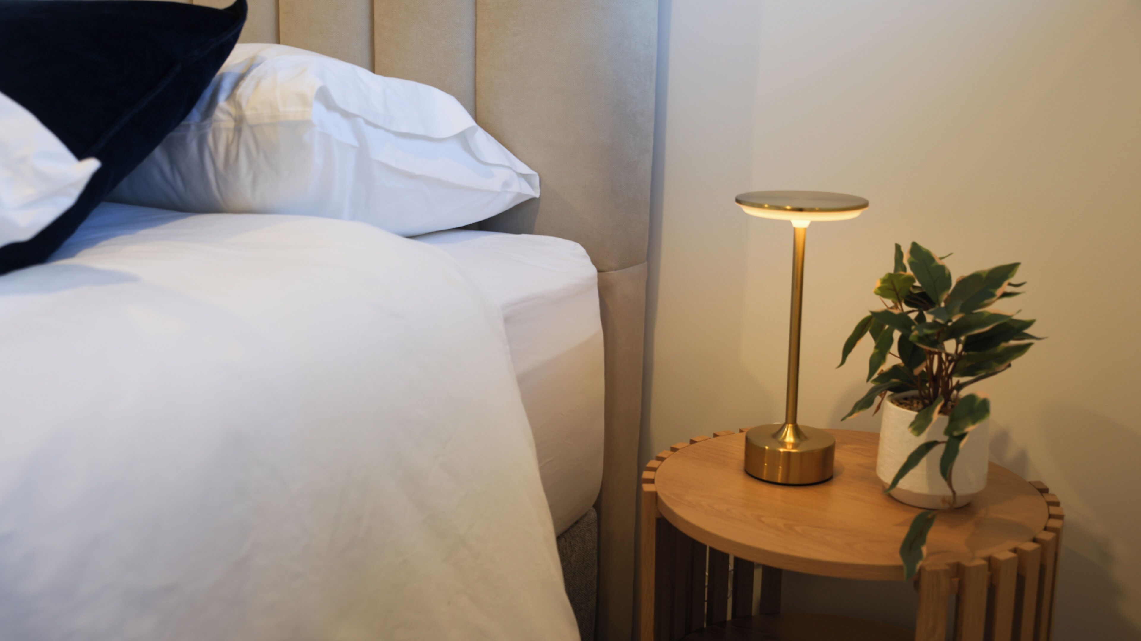 Una cama bien hecha con sábanas blancas, una lámpara de noche y una planta en maceta sobre una mesita de madera.