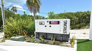 A power box labeled 'GENMAC' with a website address, surrounded by greenery and a clear sky. - Brand New Beach Cottage with Pool, Walk to La Bougainvillea Restaurant and Bar (Governor's Harbour)