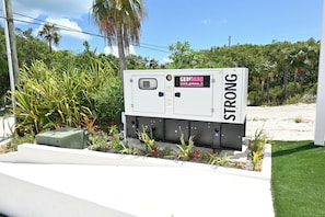 A power box labeled 'GENMAC' with a website address, surrounded by greenery and a clear sky. - Brand New Beach Cottage with Pool, Walk to La Bougainvillea Restaurant and Bar (Governor's Harbour)