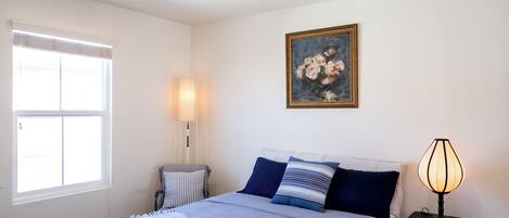 Une chambre à coucher avec un lit, une lampe de chevet, une fenêtre avec des stores et un tableau au mur.