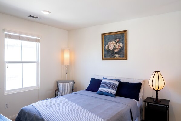 A bedroom with a bed, a bedside lamp, a window with blinds, and a painting on the wall.