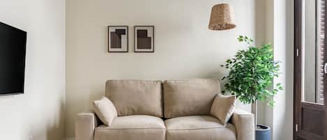 Een woonkamer met een beige bank, een witte bijzettafel, een hangende geweven lamp en een pot met een plant.