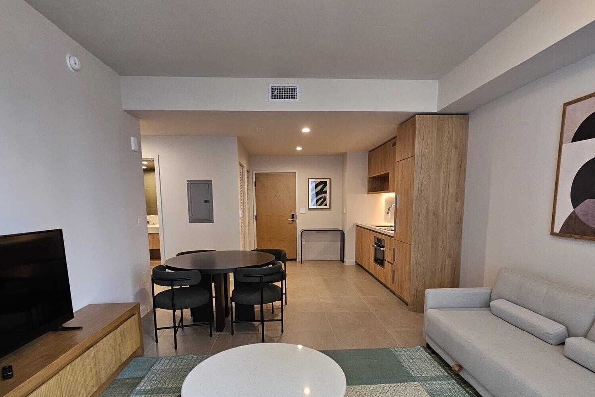 A modern living room with a sofa, a TV, a dining table with chairs, and a kitchen area in the background.