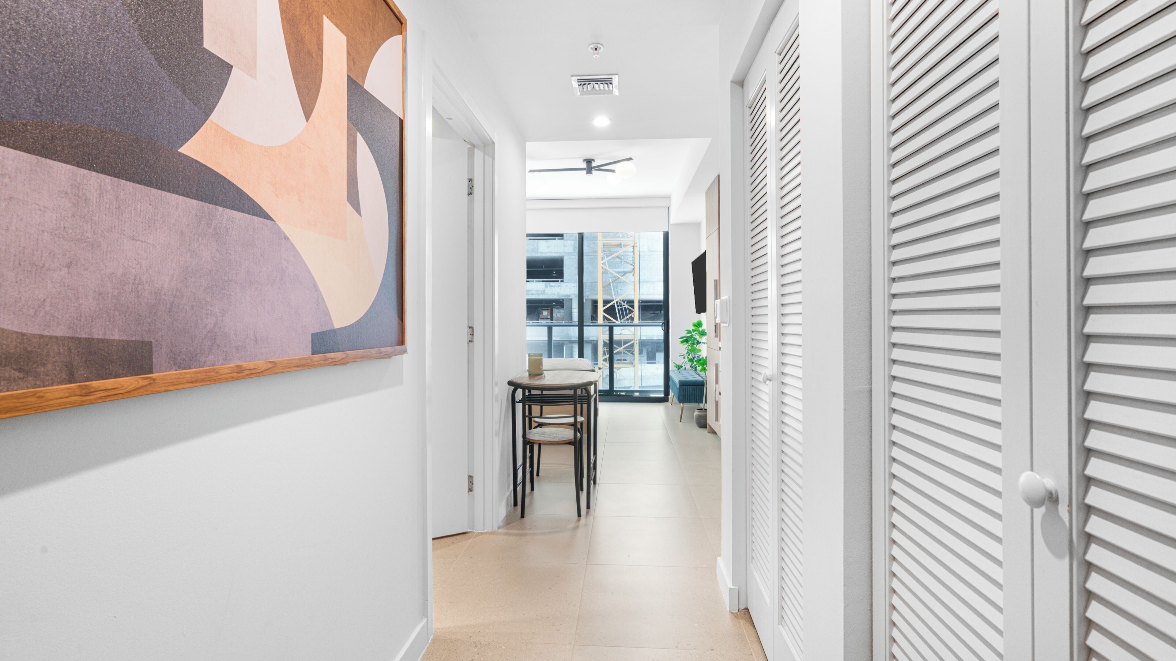 A hallway with a ceiling fan, a large abstract painting, a table with chairs, and a view of the outside through a window.
