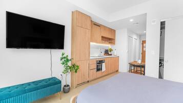 A modern bedroom with a bed, a TV, a bench, and a kitchen area with wooden cabinets and a sink.