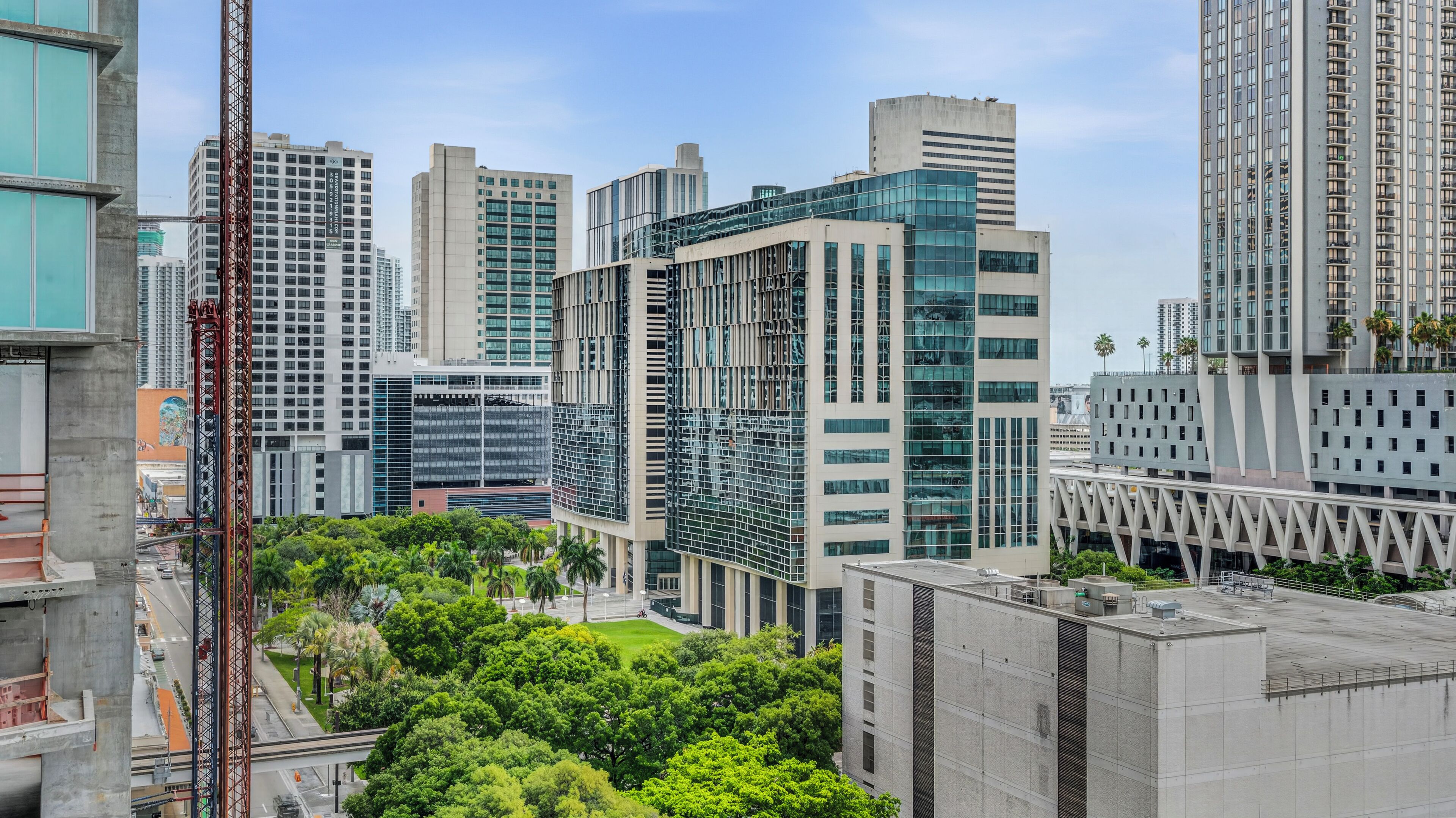 A modern urban skyline with high-rise buildings, a construction crane, and lush green trees.