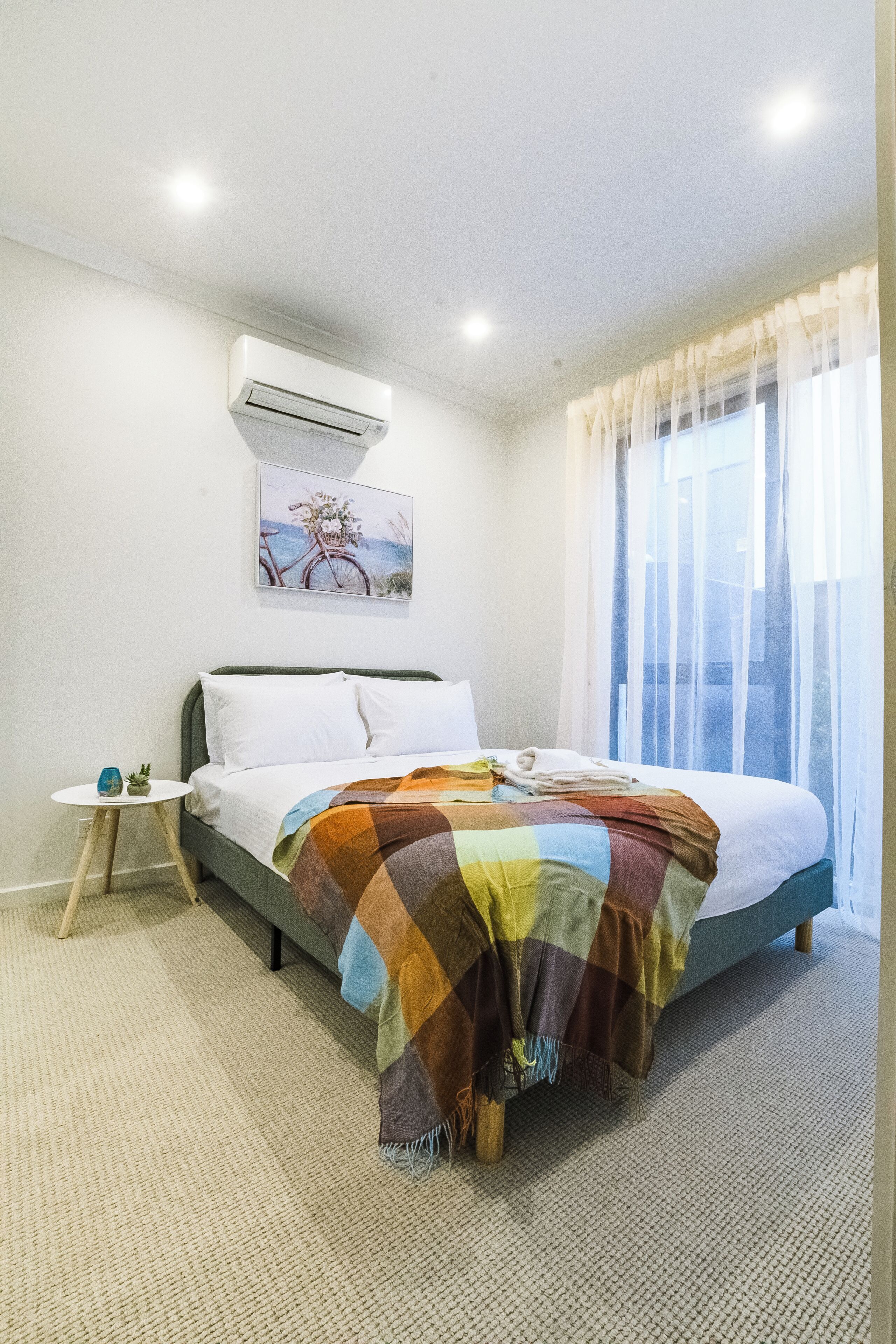 A hotel room with a bed, a small table with a vase and plant, an air conditioning unit, and a window with sheer curtains.