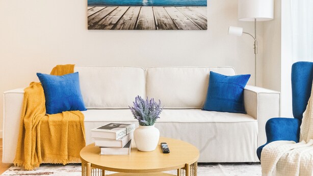 Ein Wohnzimmer mit einem weißen Sofa, blauen Kissen, einem Holztisch und einem Bild von einem Segelboot an einem Steg.