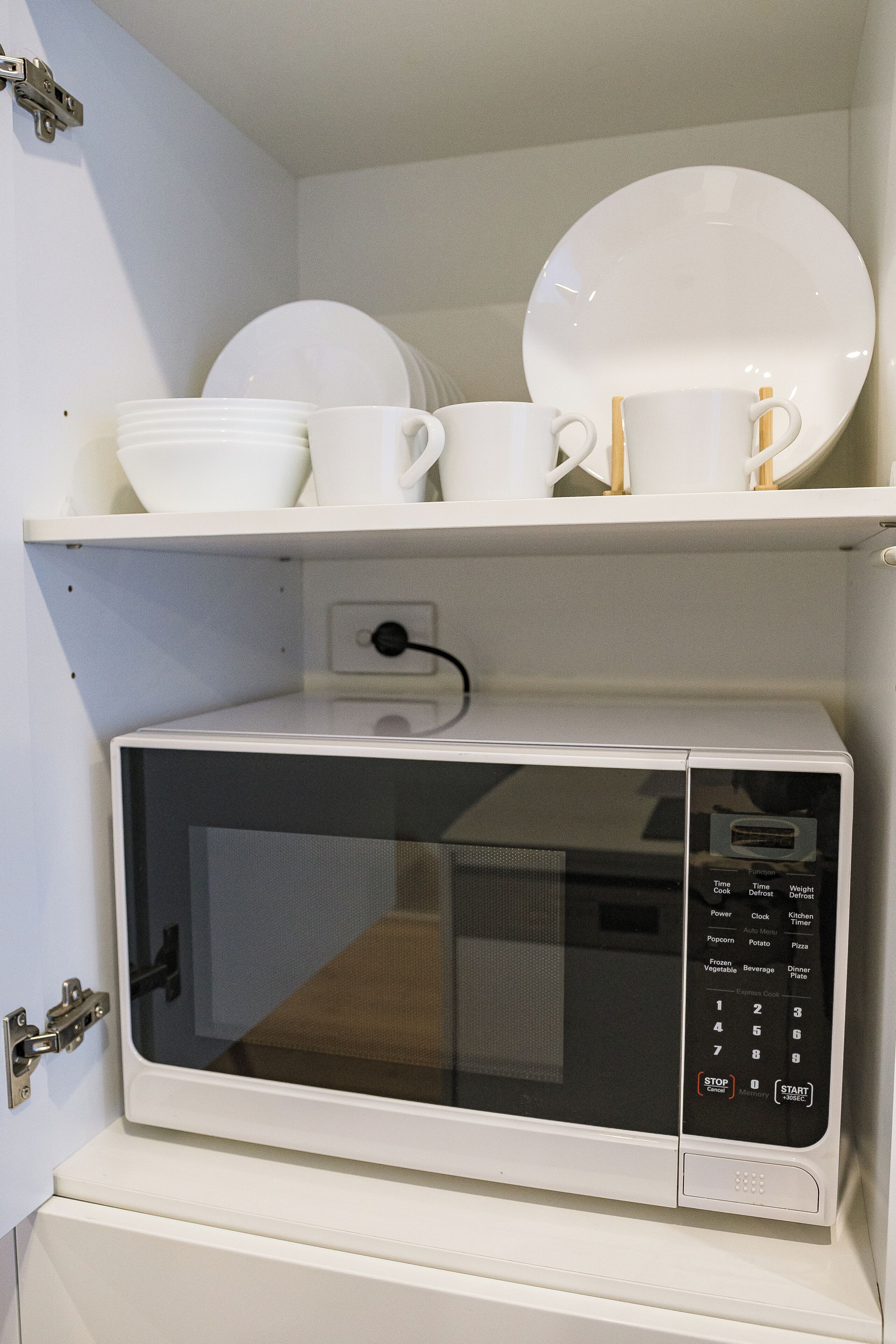 A microwave oven with a digital control panel and a shelf above it holding white dishes and cups.