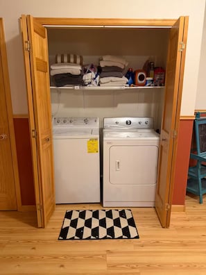 A laundry room with two washing machines, a dryer, and a shelf with folded towels and cleaning supplies. - Highwater House - Arkansas River Basecamp #020886 (Salida)