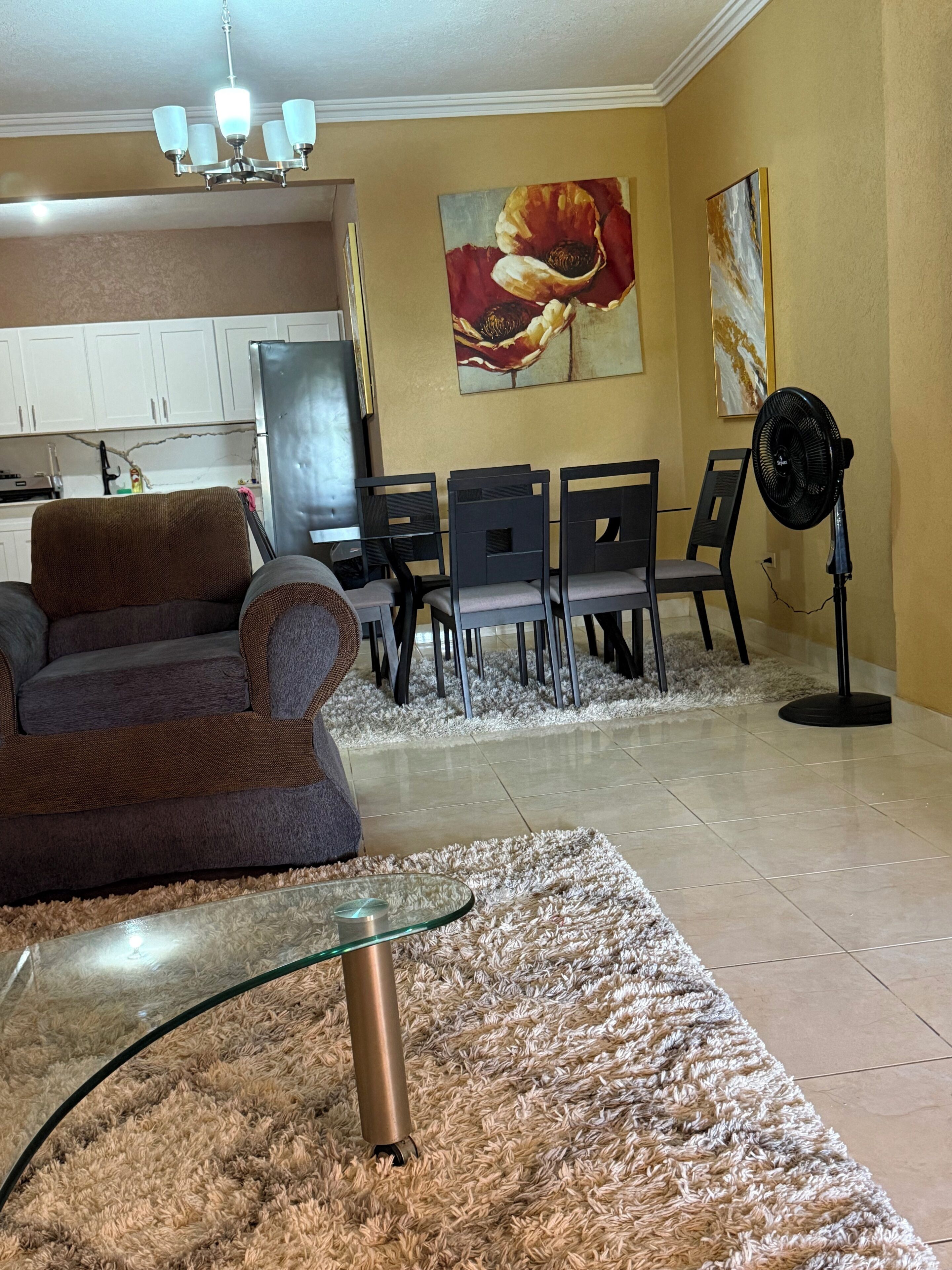 A living room with a sofa, a glass coffee table, a rug, a dining area with chairs, a fan, and artwork on the wall.