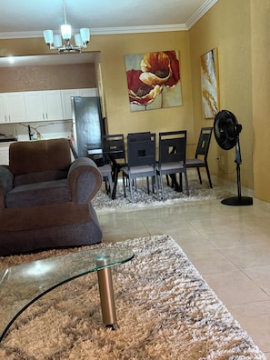 Un salon comprenant un canapé, une table basse en verre, un tapis, un coin repas avec des chaises, un ventilateur et des œuvres d’art accrochées au mur.