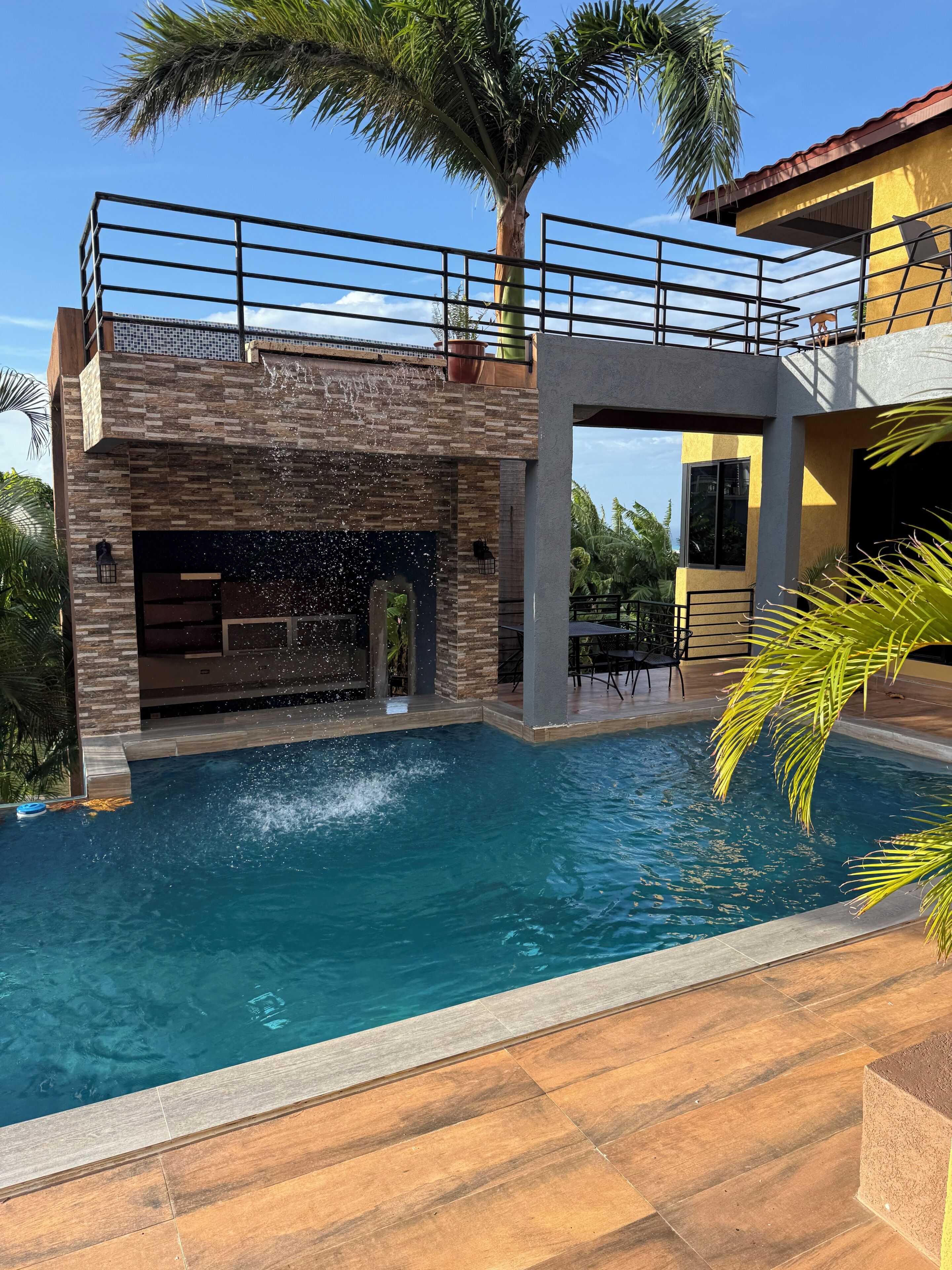 Ein Schwimmteich mit Wasserfall, umgeben von einem modernen Haus mit Balkon und einer Palme.