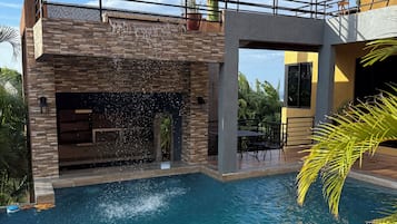 A swimming pool with a waterfall feature, surrounded by a modern house with a balcony and a palm tree.