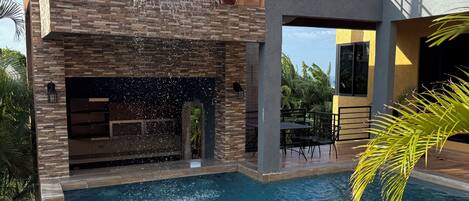 A swimming pool with a waterfall feature, surrounded by a modern house with a balcony and a palm tree.