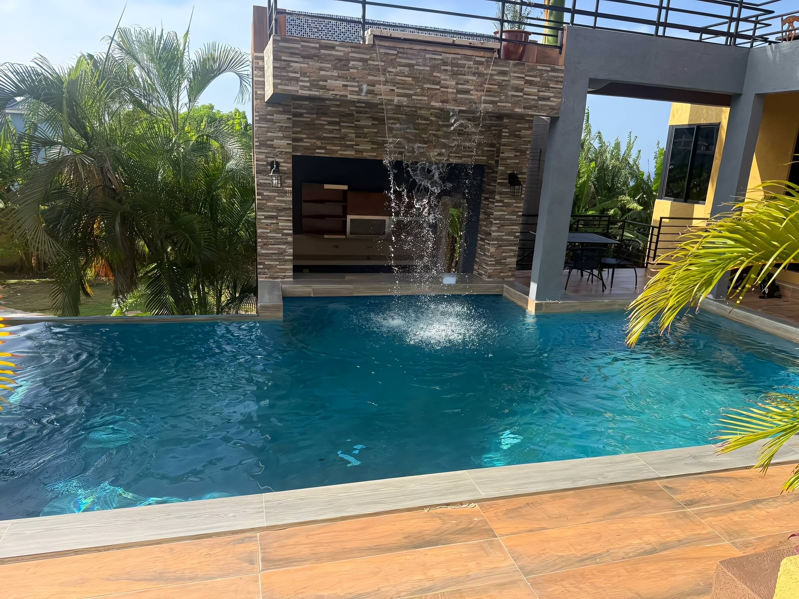 A swimming pool with a waterfall feature, surrounded by palm trees and a modern building.