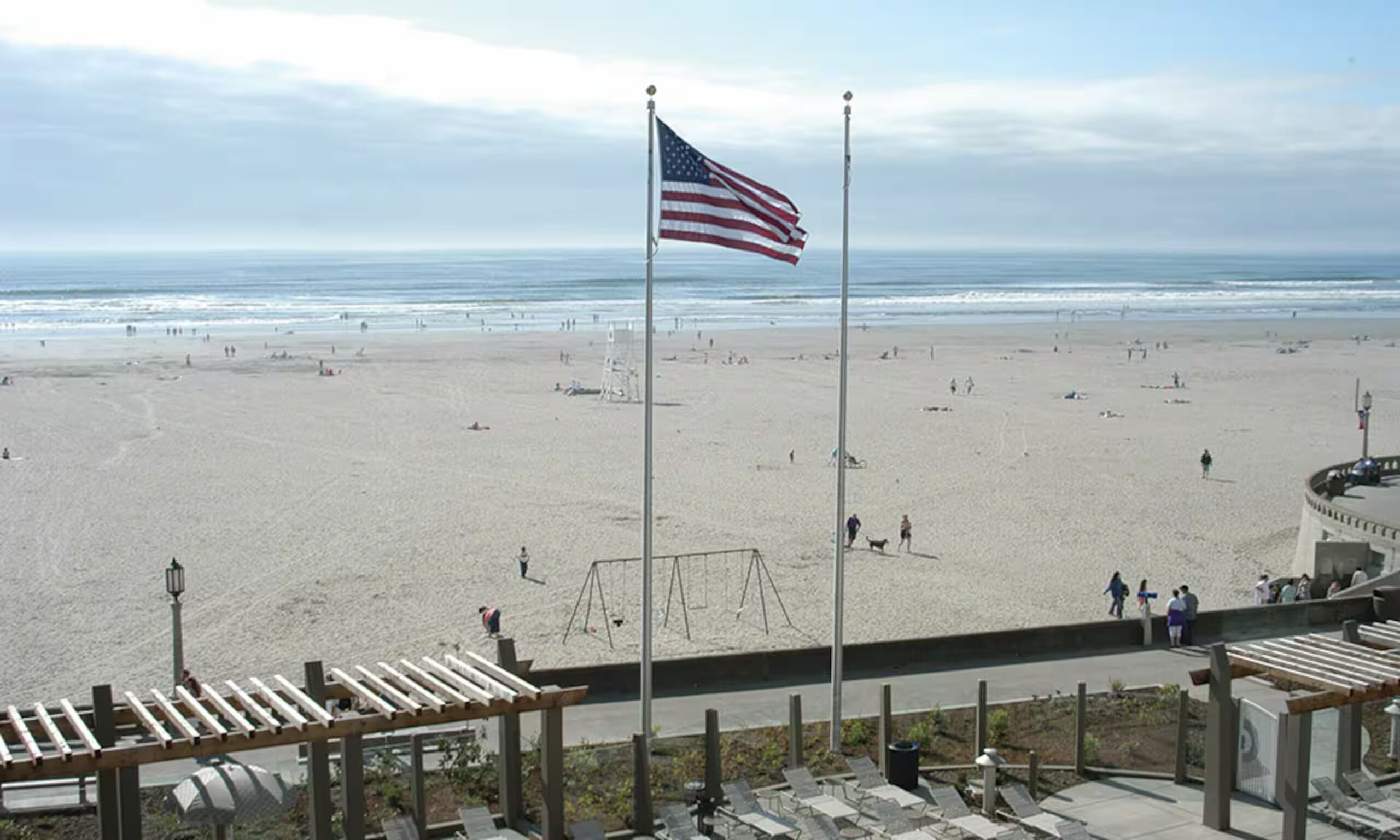 A beach with a flagpole, beachgoers, and a boardwalk with seating.